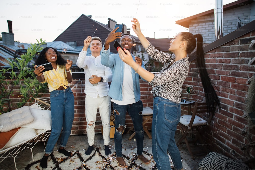 Cheerful young people in stylish clothes using smartphones and sparklers during rooftop party. Happy mixed race friends spending leisure time with fun.