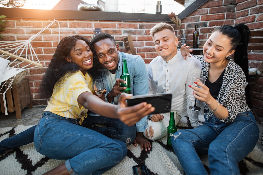 Cheerful young men and women taking selfie on modern smartphone while relaxing together on open terrace. Multicultural friends spending party time with tasty snacks and alcoholic drinks.