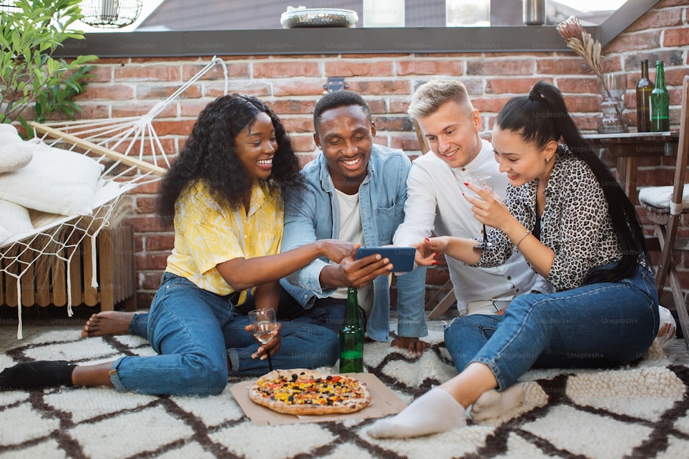 다민족 친구들이 술과 피자를 들고 열린 테라스에 앉아 현대 스마트폰으로 재미있는 비디오를 보고 있습니다. 파티, 기술 및 휴식의 개념.