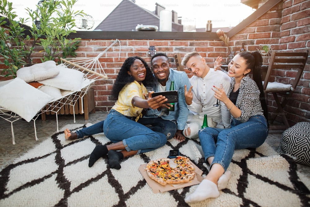 Fröhliche junge Männer und Frauen machen Selfies auf modernem Smartphone, während sie gemeinsam auf der offenen Terrasse entspannen. Multikulturelle Freunde verbringen Partyzeit mit leckeren Snacks und alkoholischen Getränken.