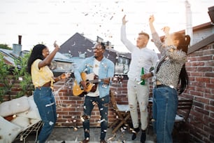 Group of four multiracial people in casual clothes drinking alcohol, playing guitar, singing and throwing up confetti. Concept of party time and enjoyment.