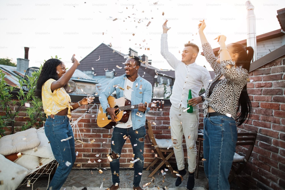 캐주얼 한 옷을 입은 4 명의 다인종 사람들이 술을 마시고, 기타를 연주하고, 노래하고, 색종이를 던지고 있습니다. 파티 시간과 즐거움의 개념.