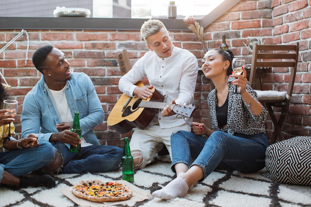 Gruppo multietnico di persone che suonano la chitarra e cantano durante la festa sul tetto. Giovani hipster che bevono alcolici e mangiano pizza. Tempo di festa durante i fine settimana.