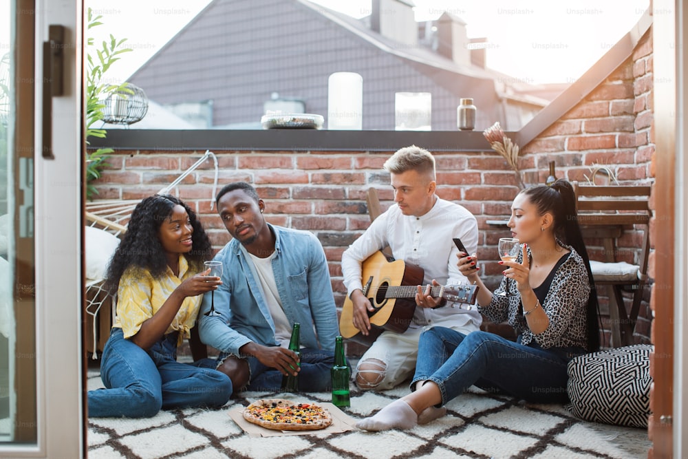 Vier multirassische Freunde sitzen auf offener Terrasse, essen leckere Pizza und trinken Alkohol. Hübscher Mann, der Gitarre spielt und singt. Partyzeit.