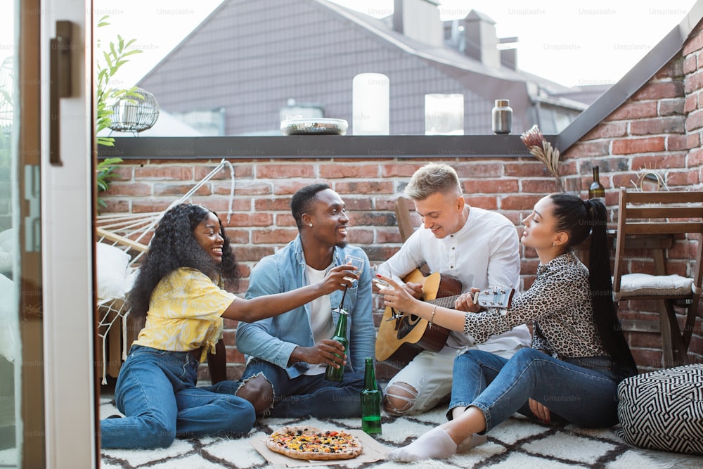 Group of diverse young people sitting together on rooftop with cocktails and pizza. Cheerful friends using guitar for relaxation during free time.