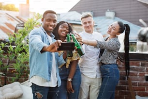 Alegres amigos mestiços brindando com bebidas alcoólicas e tirando selfies no smartphone moderno. Jovens do sexo masculino e feminino tendo tempo de festa com diversão.