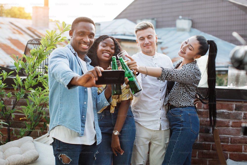 Cheerful mixed race friends toasting with alcoholic drinks and taking selfie on modern smartphone. Young male and female people having party time with fun.