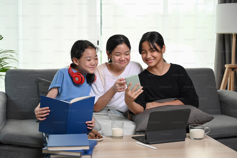 Three young girls watching something funny on smart phone while siting together on couch in living room.