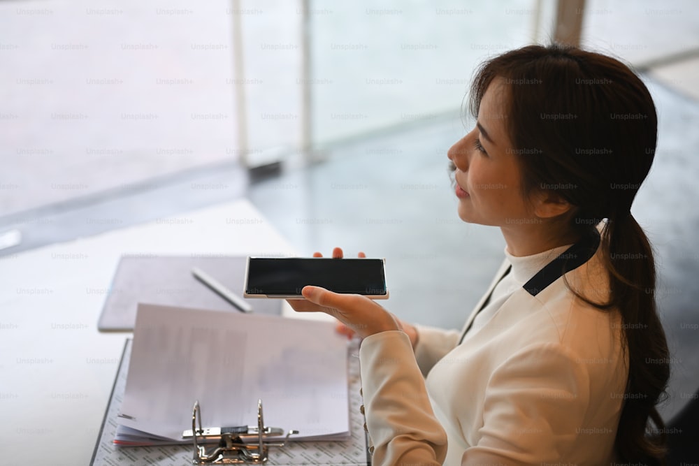 Successful businesswoman sitting in modern workplace and talking on speakerphone.