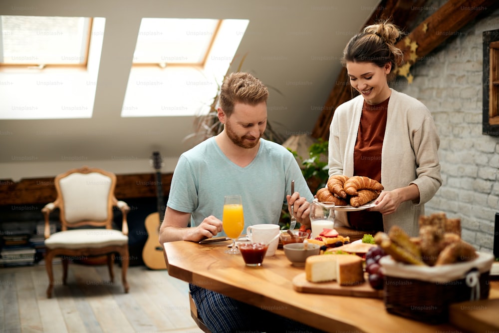 아침에 식탁에서 아침을 먹는 젊은 부부. 행복한 여자가 테이블에서 크로와상을 제공하고 있습니다.