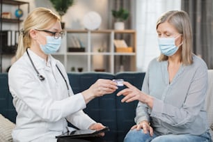 Blonde Ärztin in medizinischem Kittel, Brille und Schutzmaske, die Sauerstoffsättigung und Herzfrequenz mit einem Pulsoximeter misst, während sie ihre kranke Rentnerin zu Hause besucht.
