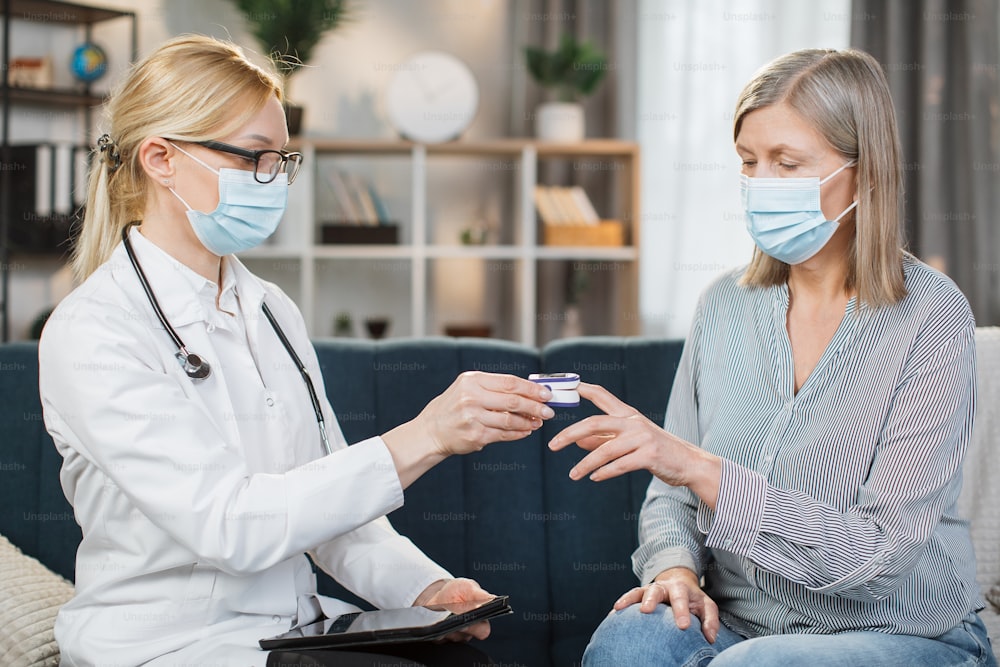 Blond female doctor in medical coat, glasses and protective mask, measuring oxygen saturation and heart rate with a pulse oximeter, while visiting her sick retired lady patient at home.