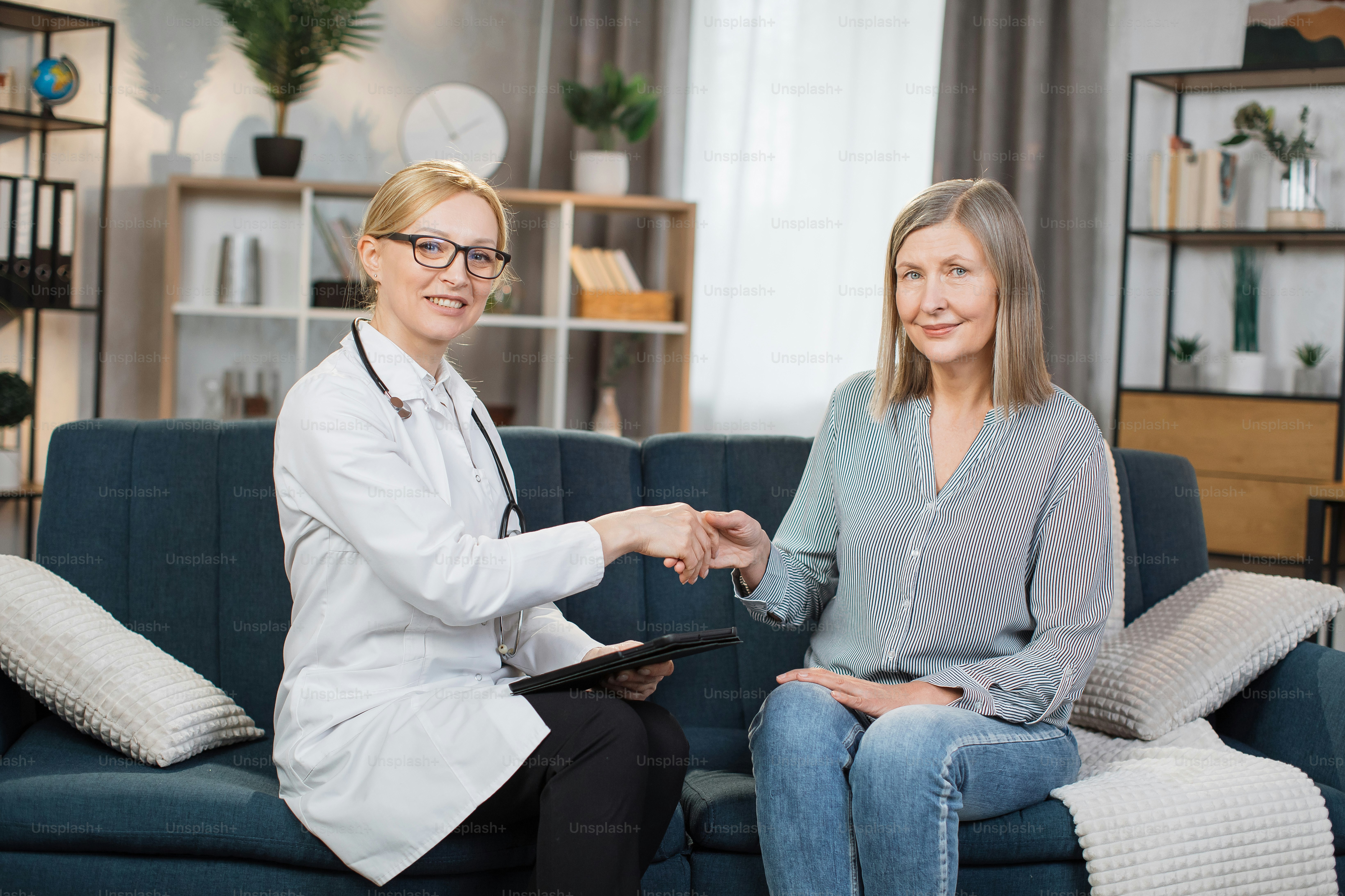 Front view of good-looking professional female Caucasian doctor in medical coat, consulting her senior patient and handshaking after sighning an insurance, during home visit.