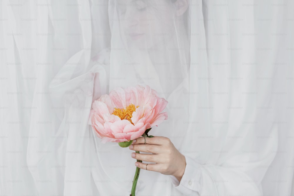 Sensual beautiful woman behind soft white fabric with pink peony in hands. Young stylish female gently holding big pink peony flower. Tender image. Spring aesthetics. Bridal morning