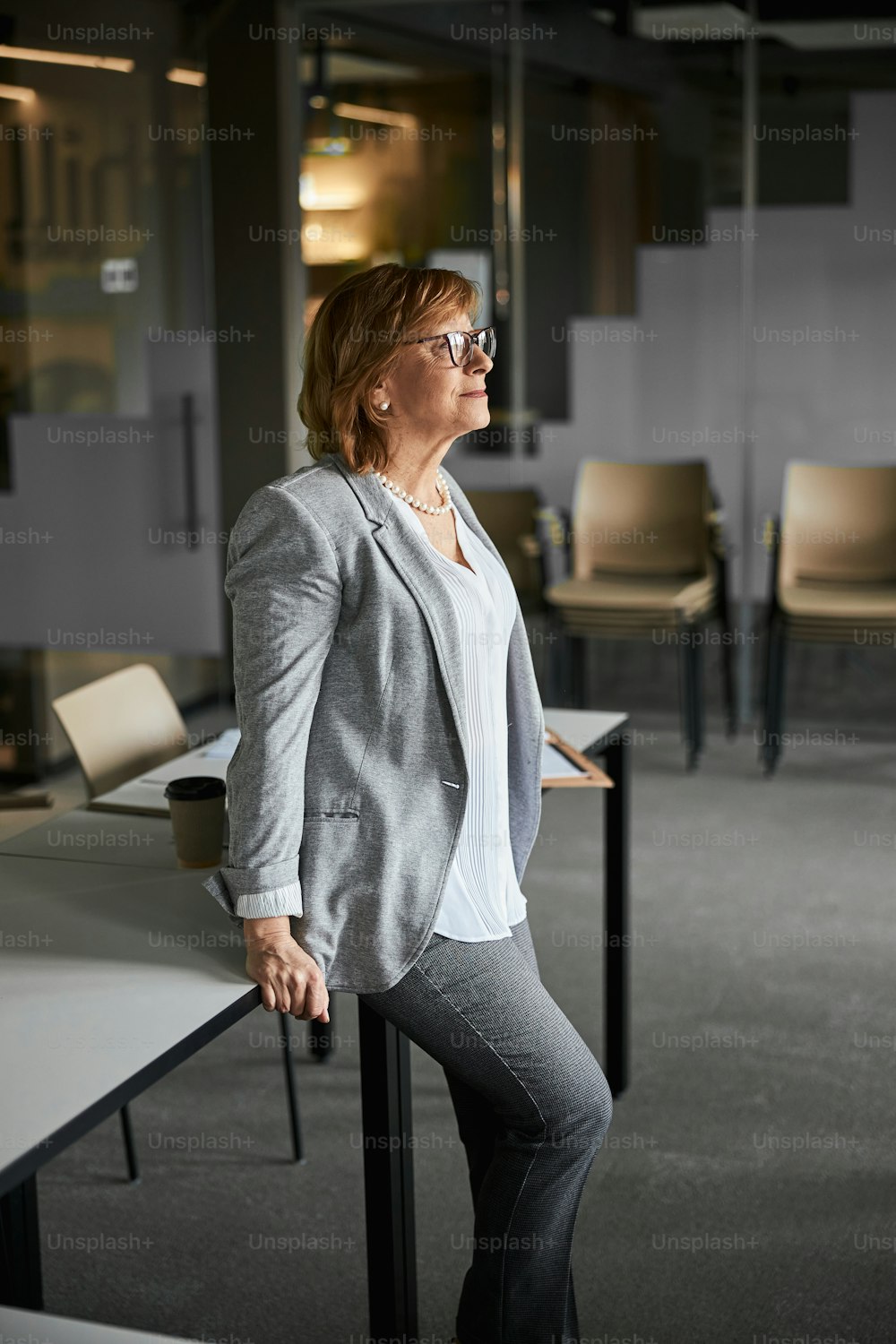 Business lady in stylish glasses and official clothing resting against a grey desk while looking ahead