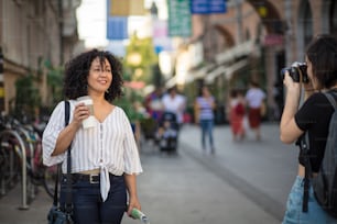 Ottima foto.  Donna sorridente in piedi sulla strada con tazza di caffè. Donna che le scatta una foto.