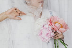 Sensual beautiful woman under soft tulle fabric holding peony bouquet in hands. Young stylish female gently holding big peonies flowers. Tender soft image. Spring aesthetics