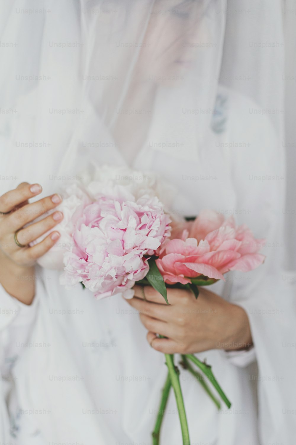 Mulher elegante bonita atrás do tecido branco macio segurando peônia nas mãos. Fêmea jovem segurando suavemente grandes flores de peônias cor-de-rosa. Imagem suave sensual. Estética primaveril. Dia da Mulher