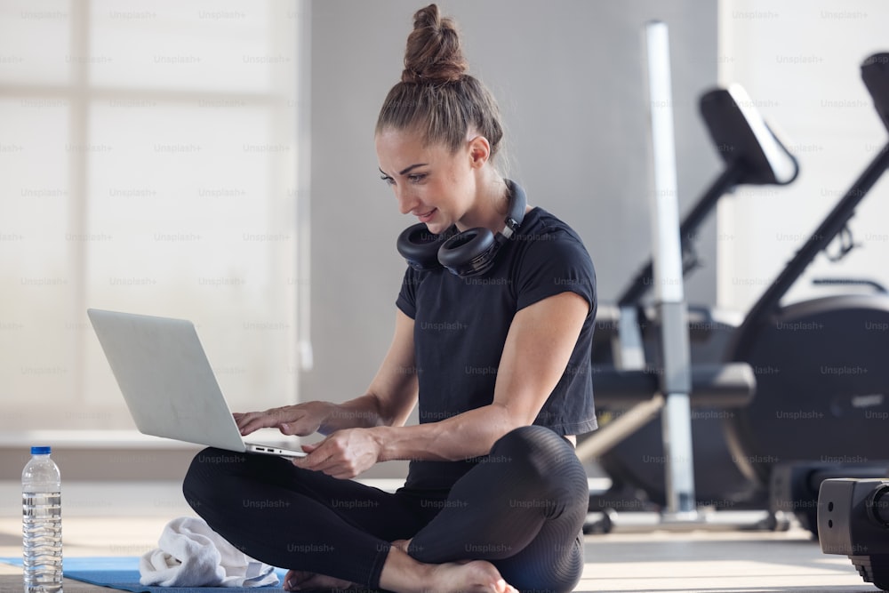 Une femme sportive en tenue de sport est assise sur le sol avec des haltères et un shake protéiné ou une bouteille d’eau et utilise un ordinateur portable à la maison dans le salon. Concept de sport et de loisirs.