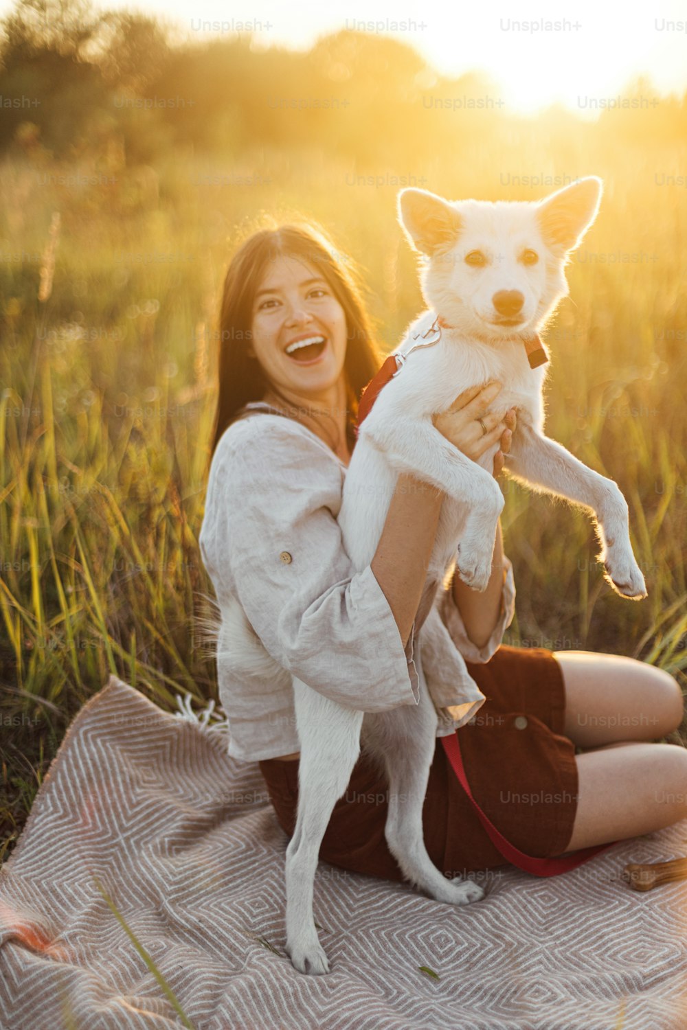 Stylish woman playing with her white dog on blanket in warm sunny light in summer meadow. Summer vacation and picnic with pet. Young boho female having fun with swiss shepherd puppy in sunset