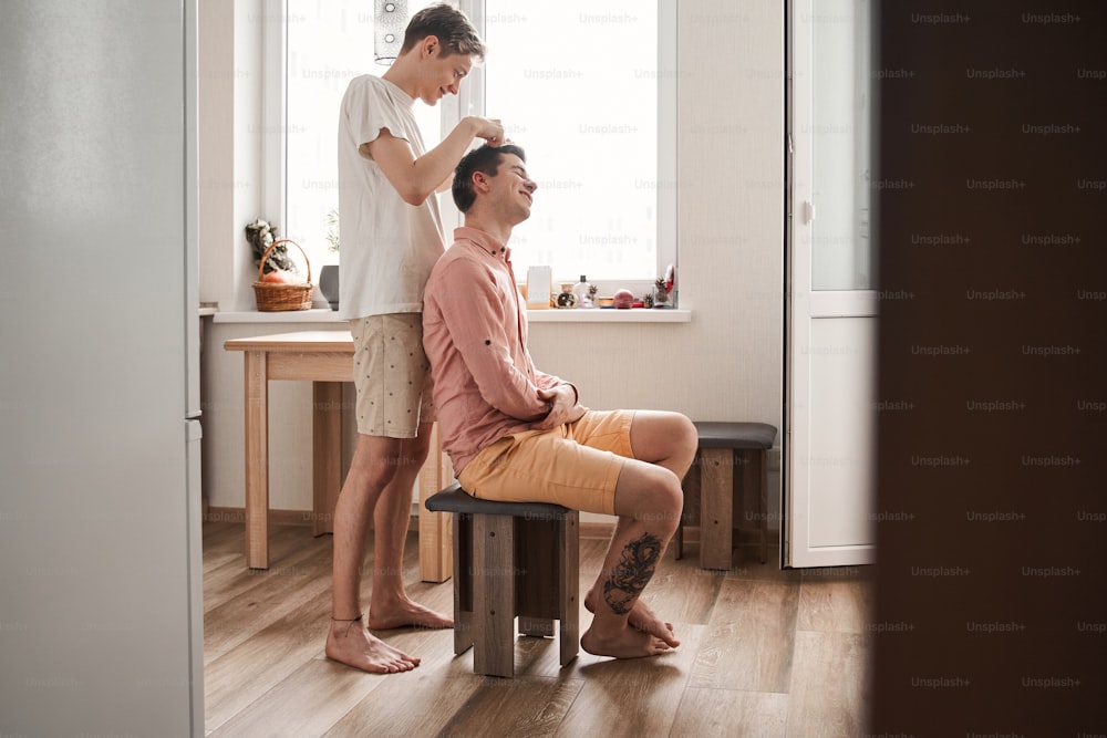 新しい標準。コロナウイルスcovid-19パンデミック中にボーイフレンドによって自宅で散髪されている白人男性の全身写真。同性愛関係と権利の概念。ストックフォト