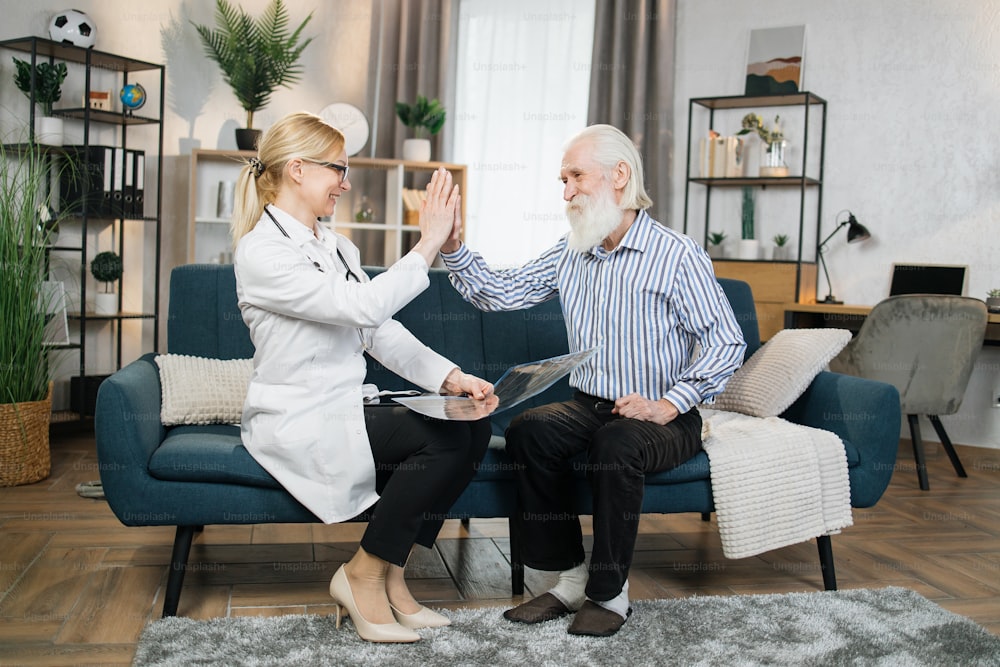 Cheerful positive blond woman doctor satisfied from successful treatment and result of x-ray CT scan of her retired male patient and giving high five each other during home visit.