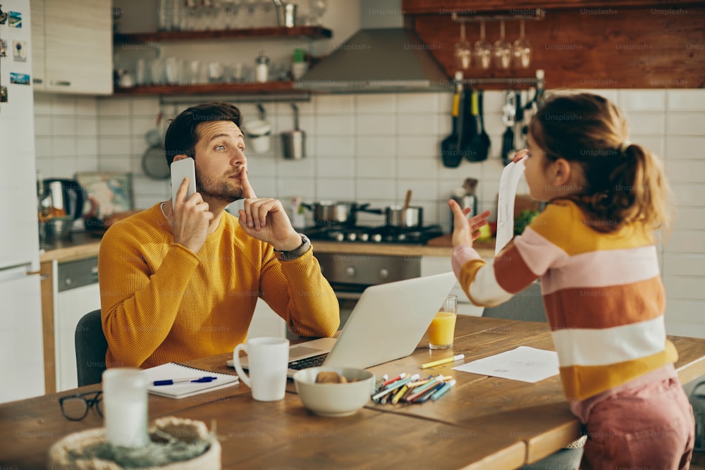 Working father talking on the phone and silencing his daughter who is showing him her drawing at home.