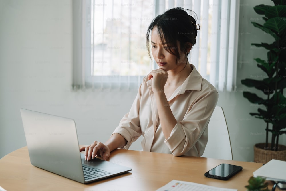 Junge asiatische Geschäftsfrau denkt über etwas nach, während sie vorne sitzt und tragbare Laptops liest E-Mails vom Kunden, langes Arbeitszeitkonzept
