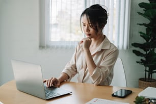 Joven empresaria asiática pensando en algo mientras está sentada frente a la computadora portátil portátil abierta leyendo el correo electrónico del cliente, largas horas de concepto de trabajo