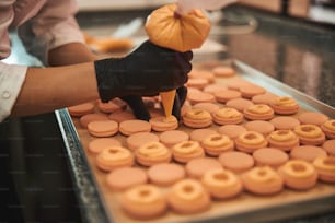 Abgeschnittenes Foto eines talentierten Konditors, der Glasur auf jedes Macaron auf dem Backblech drückt