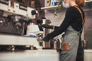 Photo recadrée d’une dame non reconnue portant un masque et un uniforme de barista debout près d’une machine à café et préparant un expresso
