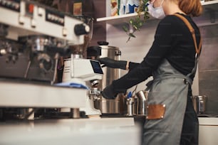 Cropped photo of unrecognised lady in mask and barista uniform standing near coffee machine and making expresso