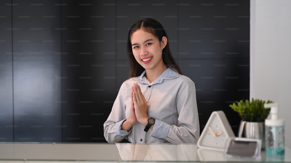 Joven recepcionista asiática sentada en el mostrador de recepción y levantando las manos para dar la bienvenida a los visitantes al hotel.