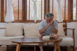 Asian senior man sitting on sofa in home living room and reading a book. Stressed elderly grandfather suffering from eye strain and headache. Retirement old people health care and eye vision problem concept.
