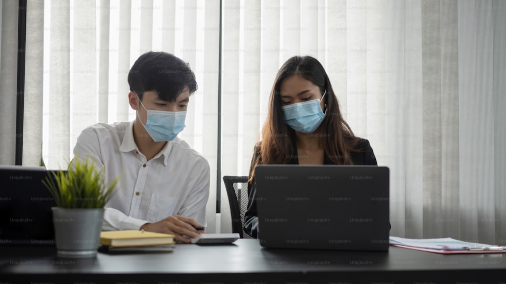 Jóvenes empresarios asiáticos con máscara protectora discutiendo ideas de negocios y trabajando juntos en la oficina.