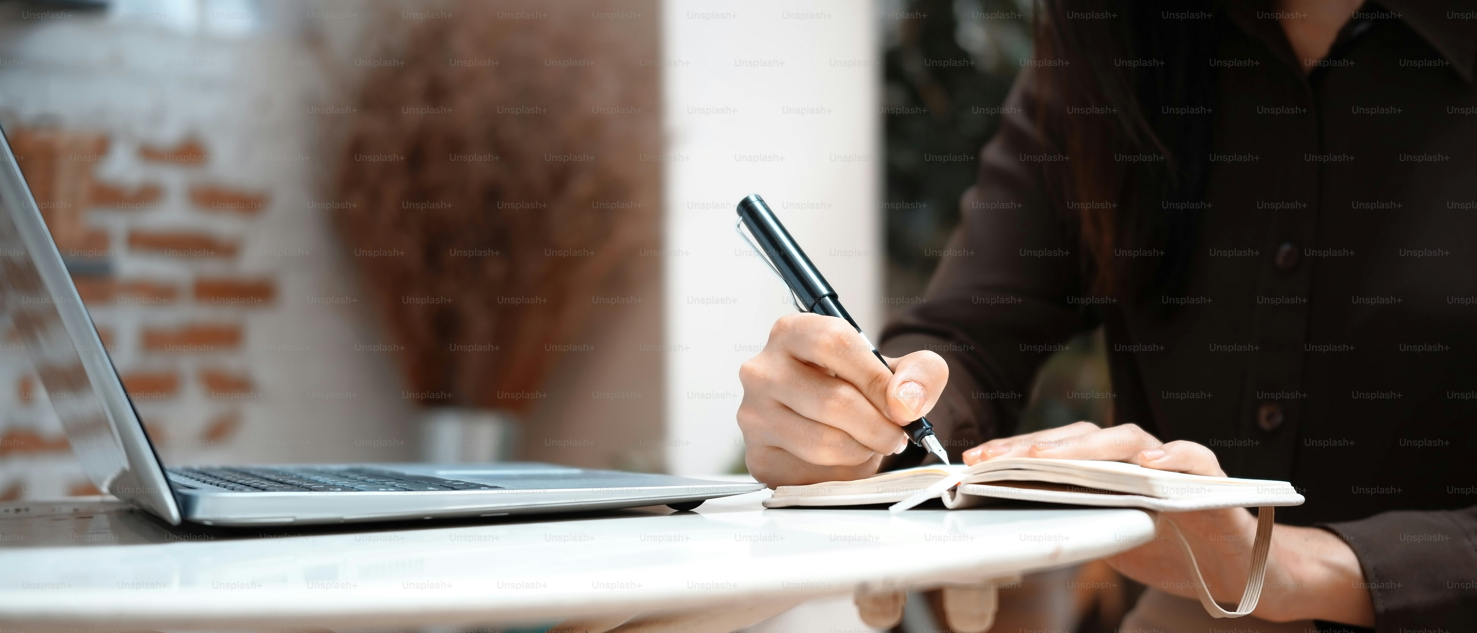 Cropped shot of young female freelancer working with laptop computer and writing notes into notebook.
