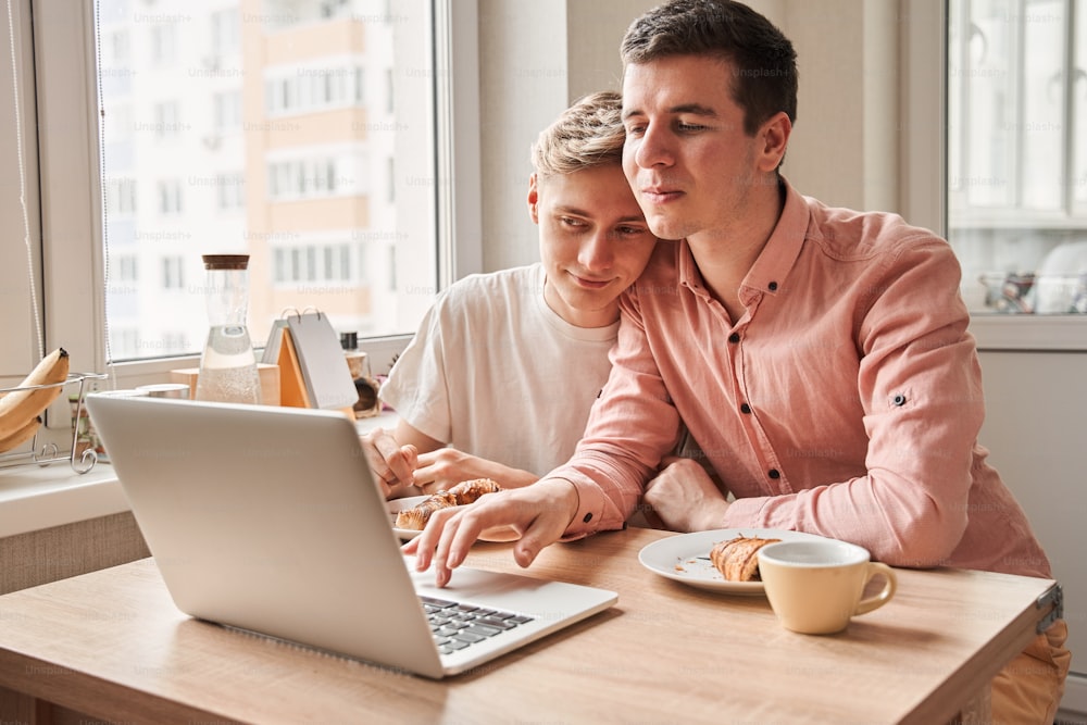 이것 보세요. 잘 생긴 게이 커플은 부엌에서 맛있는 아침 식사를 하면서 노트북으로 무언가를 보고 있다. 부부 관계 개념입니다. 스톡 사진