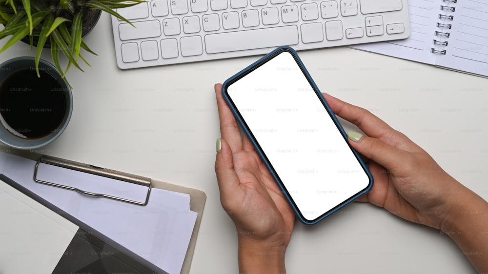 Above view of woman hands holding mobile phone with empty screen on whote office desk.