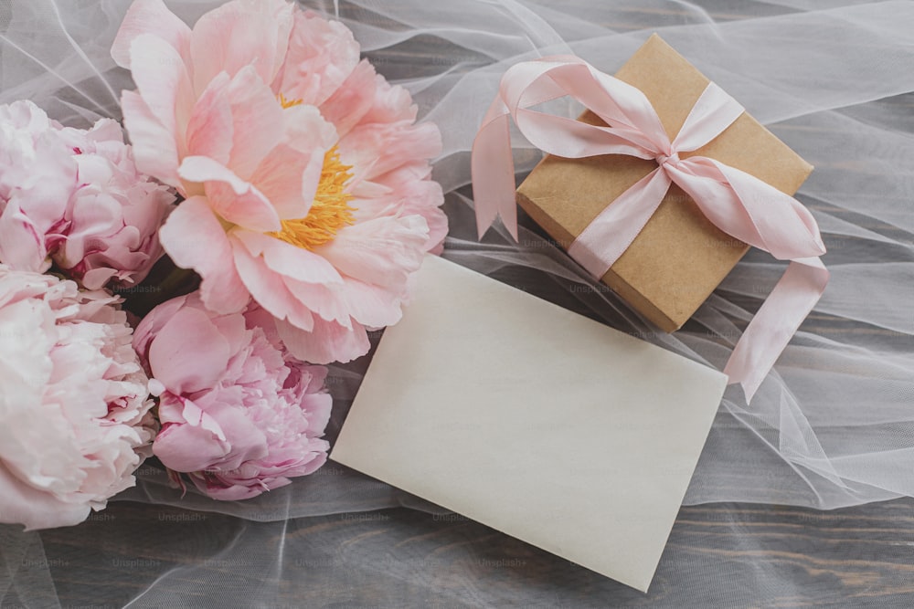 Beautiful peony bouquet, gift box and card on soft tulle fabric on dark wooden background, top view. Happy Mothers day. Greeting card template. Pink and white peonies flowers and stylish present