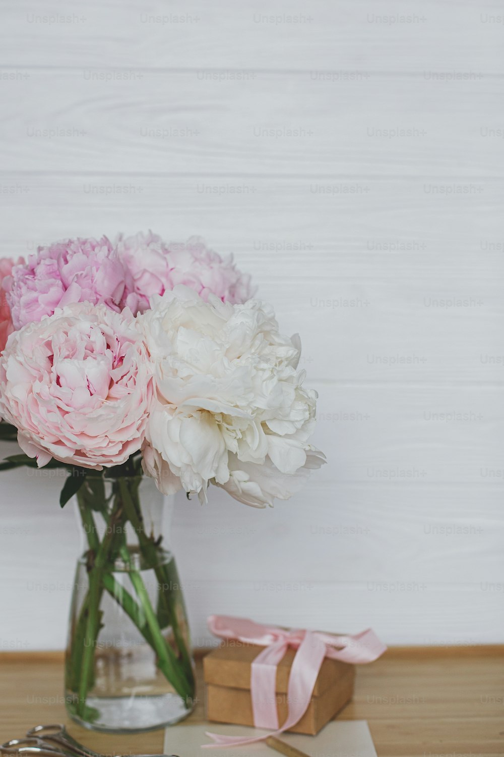 Beautiful peony bouquet in vase, gift box, greeting card and scissors on wooden background, copy space. Happy Mothers day. Pink and white peonies and present