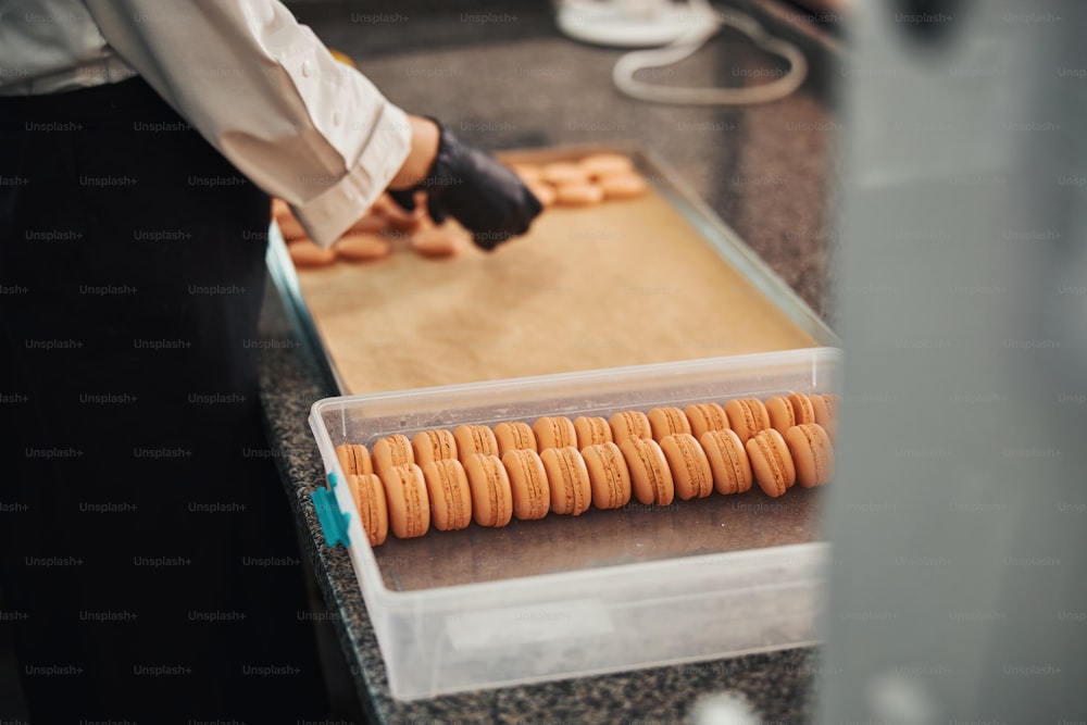 Foto recortada de un cocinero profesional clasificando y empacando innumerables macarons de color melocotón en recipientes