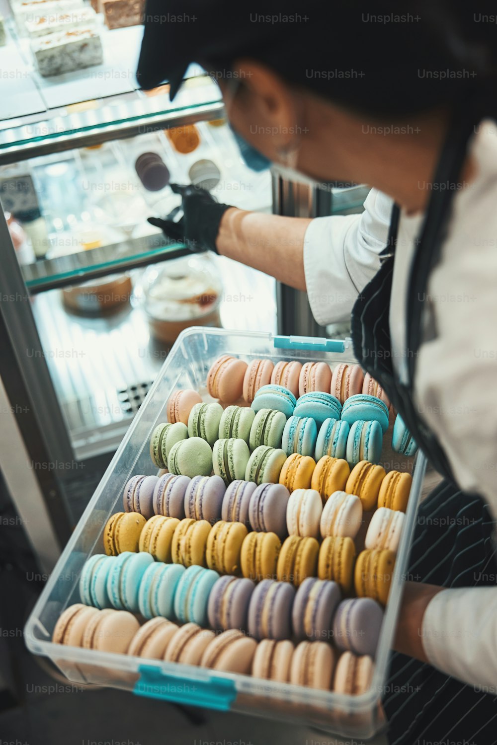 Cropped photo of cafe employee in mask holding box with macarons while adding some to the window display