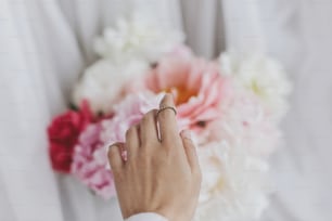 Hand with stylish rings on beautiful peonies bouquet. Big white and pink peony flowers and bride hand. Tender image. Wedding bouquet. Florist arranging flowers. Feminine essentials