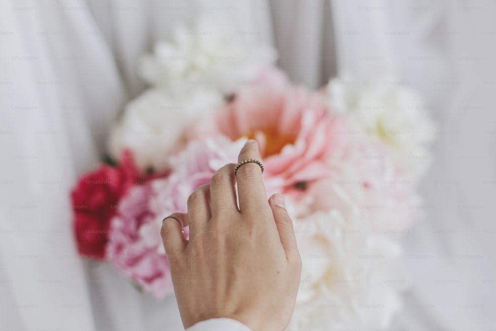 Hand with stylish rings on beautiful peonies bouquet. Big white and pink peony flowers and bride hand. Tender image. Wedding bouquet. Florist arranging flowers. Feminine essentials