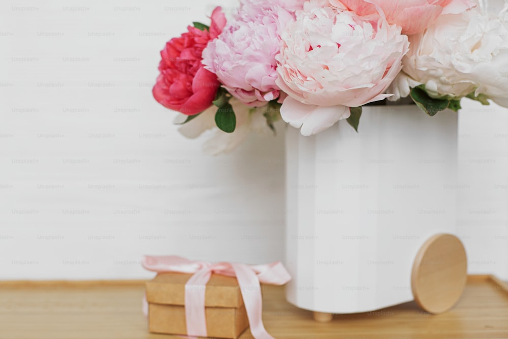 Beautiful peony bouquet in modern stylish vase and gift box on wooden background, copy space. Happy Mothers day. Pink and white peonies and present