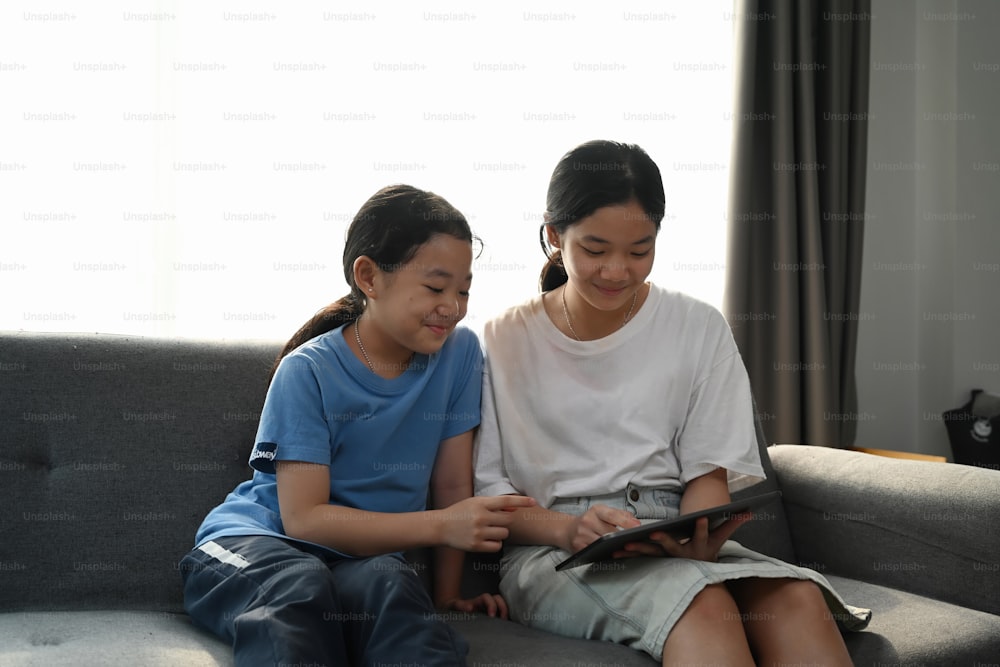 Two young girls sitting on cozy couch and watching something on digital tablet together.