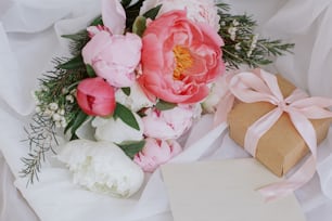 幸せな母の日。素朴な椅子の上の柔らかい白い生地の美しいスタイリッシュな牡丹の花束、グリーティングカード、ギフトボックス。ピンクと白の牡丹の花、プレゼント、ポストカード。結婚式の招待状