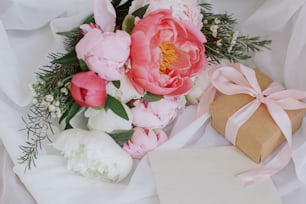 Happy Mothers day. Beautiful stylish peonies bouquet, greeting card and gift box on soft white fabric on rustic chair. Pink and white peony flowers, present, postcard. Wedding invitation