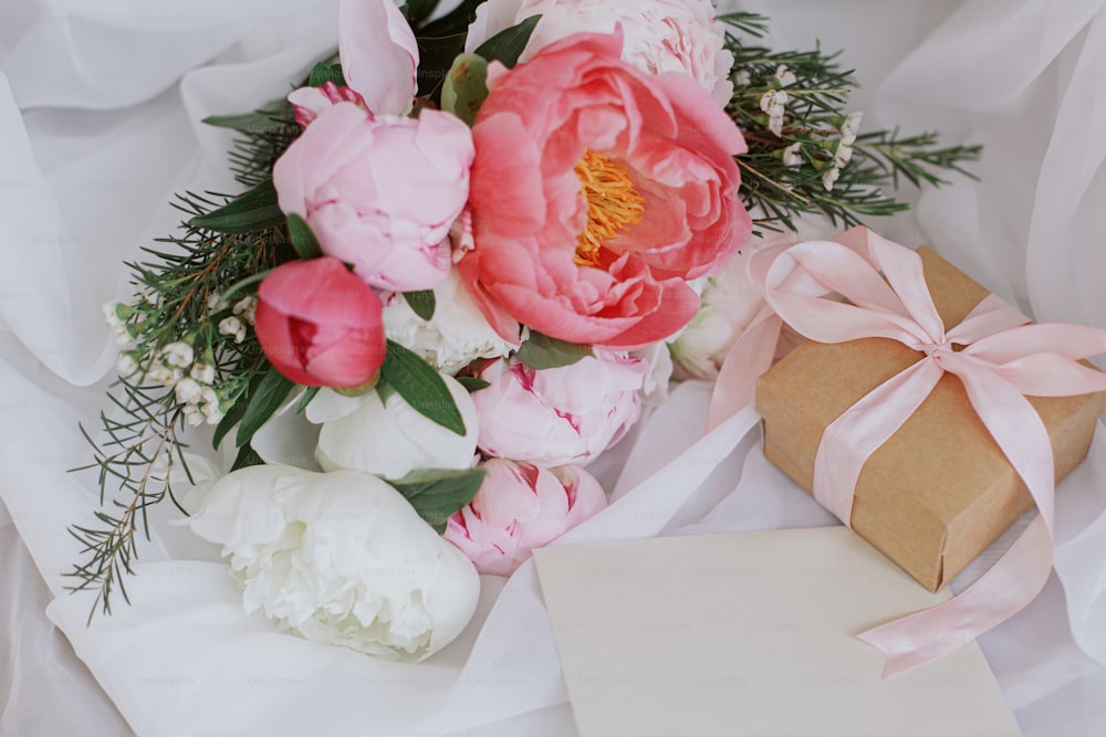 Alles Gute zum Muttertag. Schöner stilvoller Pfingstrosenstrauß, Grußkarte und Geschenkbox auf weichem weißem Stoff auf rustikalem Stuhl. Rosa und weiße Pfingstrosenblüten, Geschenk, Postkarte. Hochzeitseinladung