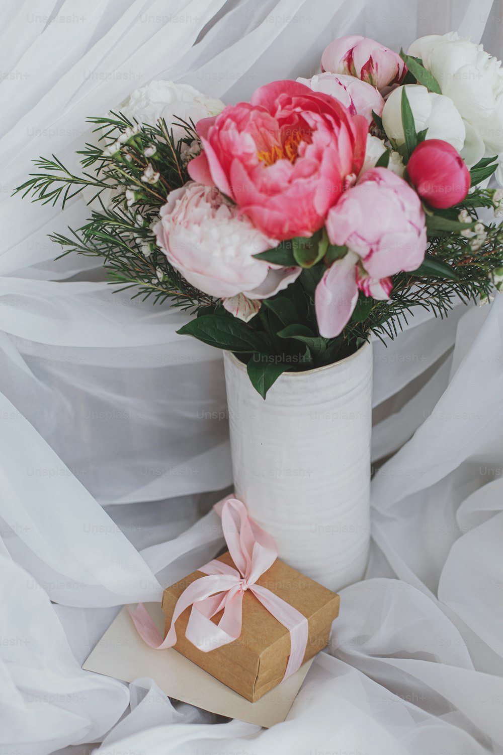 Beautiful stylish peonies bouquet, gift box and greeting card on background of soft white fabric. Pink and white peony flowers in ceramic vase. Happy Mothers day. Bridal morning